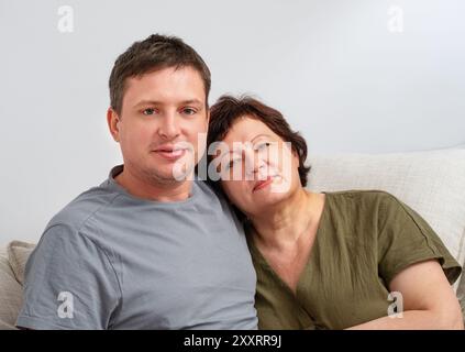 Porträt eines erwachsenen Sohnes, der seine ältere Mutter umarmt, im Wohnzimmer auf dem Sofa Stockfoto