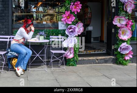 London, Großbritannien. Eine junge Frau, die vor einem Café in Camden sitzt und Kopfhörer trägt und ein Buch liest Stockfoto