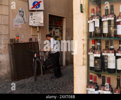 Ein Priester sperrt sein Fahrrad auf einer Kopfsteinpflasterstraße in der Nähe eines Weinladens, Campo dei Fiori, Rom, Italien. Das tägliche Leben in Rom, urbane und traditionelle Elemente Stockfoto