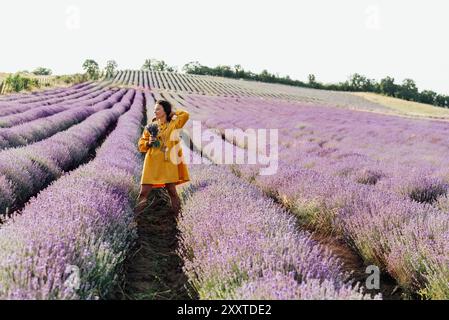 Lächelnde Frau in einem gelben Kleid mit einem Blumenstrauß aus Lavendelblumen, die auf einem malerischen Feld mit Bergen im Hintergrund steht. Stockfoto