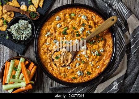 buffalo Chicken Dip in Backform auf grauen rustikalen Holztisch mit Crackern, Blaukäse, Karotten- und Selleriestangen auf Schieferbrett, horizontale Ansicht fr Stockfoto