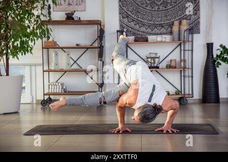 Ganzkörper eines Mannes in Sportbekleidung, der tagsüber Eka Pada Koundinyasana auf der Trainingsmatte im Zimmer macht Stockfoto