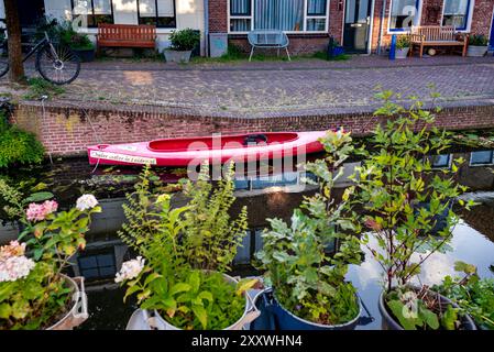 Ein rotes Kajak ruht auf einem gemauerten Gehweg neben einem ruhigen Kanal, umgeben von grünem Laub und Topfpflanzen. Fahrräder werden gegen einen Zaun geparkt, mit tr Stockfoto