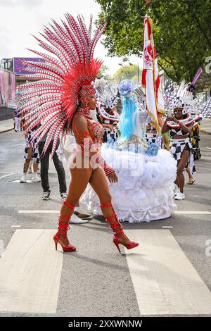 London, UK, 26. August 2024. Tänzer überqueren die Straße in Kostümen. Die Teilnehmer der Paraiso School of Samba, einer der größten Gruppen mit fast 200 Künstlern und Musikern, kommen vom frühen Morgen bis zum Beginn der Prozession um 12 Uhr. Am Montag findet der Karnevalszug von MAS-Bands, Schlagzeugern und karibischen und südamerikanischen Salsa-, Samba- und Soca-Gruppen statt. Am zweiten von zwei Tagen des Karnevalsfestes in Notting Hill feiern die Nachtschwärmer das Wochenende der Bank Holiday, an den Soundsystemen, an den Ständen und an den Veranstaltungsorten entlang der Karnevalsroute. Stockfoto