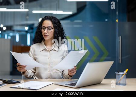 Geschäftsfrau, die Papiere am Schreibtisch im modernen Büro prüft. Professionelle Frau, die tief nachdenkt und Dokumente analysiert, mit Laptop und elegantem Büro aus Glas. Stockfoto