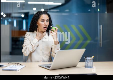Büroangestellte leiden unter Halsbeschwerden, während sie Spray zur Linderung in modernen Büroumgebungen verwenden. Frau, die Kehle hält und einen Ausdruck des Unbehagens zeigt, am Schreibtisch mit Laptop sitzt Stockfoto