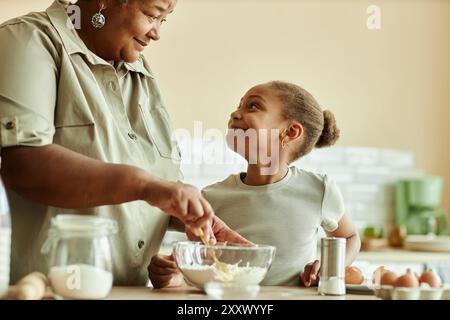 Seitenansicht des bezaubernden Schwarzen Mädchens, das die liebevolle Oma mit einem liebevollen Lächeln ansieht, während er zusammen Pfannkuchen in der beigefarbenen Küche kocht, Kopierraum Stockfoto