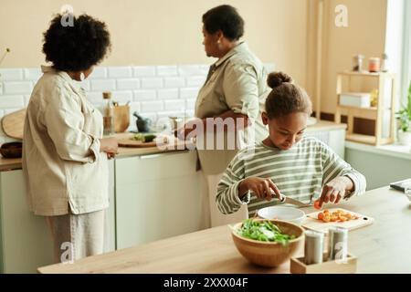 Mittelgroßer Schuss von afroamerikanischem Mädchen an der Holztheke sorgfältig schneiden Kirschtomaten für die Zubereitung von frischem Salat, während das Kochen gesundes Abendessen zusammen mit der Familie in der beigen Küche, Kopierraum Stockfoto