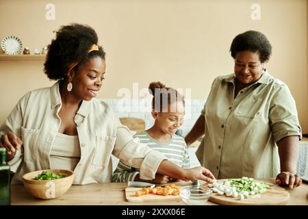 Mittlere Aufnahme der Familie Happy Black mit lächelnder Mutter, Mädchen und Großmutter, die zusammen lachen, während sie gesunden Salat für das Abendessen in der beigen Küche kochen Stockfoto