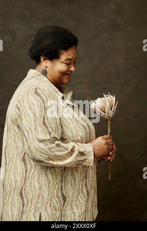 Seitenansicht der lächelnden älteren Schwarzen Frau, die sanft erhaltene König-Protea-Blume hält, die die Erhaltung von Erinnerungen und Erbe auf braunem Hintergrund im Studio, Kopierraum symbolisiert Stockfoto