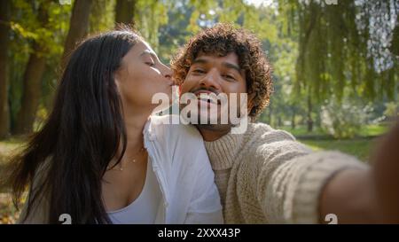 Sorgloses multirassisches Paar arabisch indischer männlicher Freund glückliche hispanische Freundin Frau macht Foto auf Handy-Gerät draußen im Park Mann Stockfoto