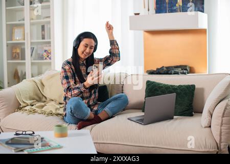 Lächelnde junge asiatische Studentin der Generation Z in Freizeitkleidung in kabellosen Kopfhörern mit einem modernen Smartphone im komfortablen Wohnzimmer Stockfoto
