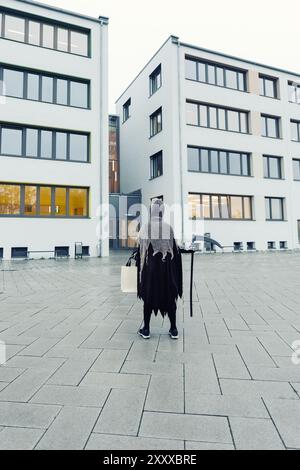 Rückansicht eines Kindes im grimmigen Sensenmann Halloween Kostüm mit Sense und Tasche für Süßigkeiten an der Fassade des Schulgebäudes bereit für die Halloween-Party Stockfoto