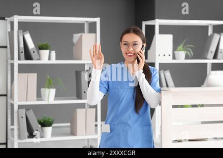 Junge Ärztin in Brille, die mit Handy spricht und OK-Geste im Krankenhaus zeigt Stockfoto