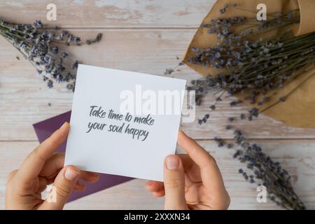 Weibliche Hände, die Papierkarten mit Text nehmen, NEHMEN SICH ZEIT, UM IHRE SEELE mit violettem Umschlag GLÜCKLICH ZU MACHEN. Lavendelblume. Draufsicht, flach. Konzept der psychisch-spirituellen Gesundheit Selbstversorgung Wohlbefinden Achtsamkeit Stockfoto
