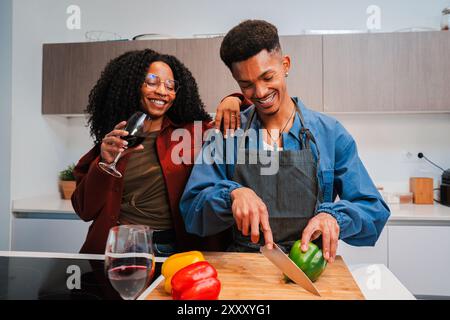 Das afroamerikanische Paar kocht veganes Essen in der Wohnküche und genießt dabei ein Glas Rotwein. Zwei junge Menschen schneiden Gemüse, um zu Hause eine gesunde vegetarische Mahlzeit zuzubereiten. Hochwertige Fotos Stockfoto