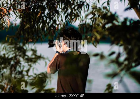 Mann, der seine Kamera auf den See zeigt. Ein Mann, der Fotos mit der Kamera am Ufer des Sees in der Abenddämmerung macht Stockfoto