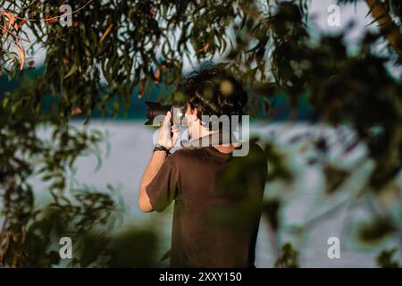 Der Mann zeigt seine Kamera mit der Hand in die Nähe des Sees. Ein Mann, der Fotos mit der Kamera am Ufer des Sees in der Abenddämmerung macht Stockfoto