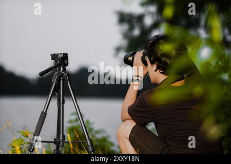 Junger Mann, der am Ufer des Sees eine Kamera aufstellt. Rückansicht der Kamera mit Stativ für männliche Einstellungen Stockfoto