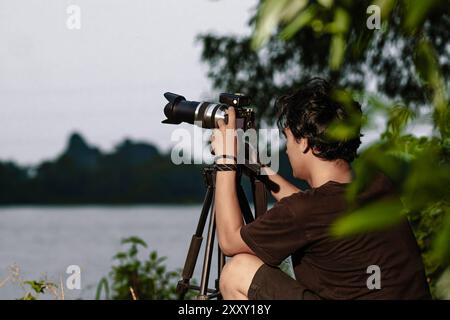 Junger Mann, der am Ufer des Sees eine Kamera aufstellt. Rückansicht des Mannes, der die Kamera mit Stativ einrichtet Stockfoto