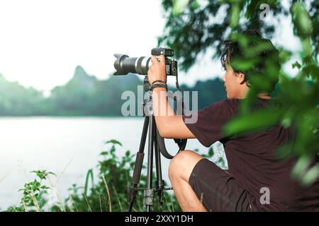 Der Mann versucht, Fotos in der Nähe des Sees mit seiner Kamera zu machen. Mann, der Fotos mit Kamera auf dem Stativ in der Nähe des Sees macht. Foto von der Rückseite Stockfoto