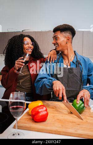 Vertikal. Das afroamerikanische Paar kocht veganes Essen in der Wohnküche und genießt dabei ein Glas Rotwein. Zwei junge Menschen schneiden Gemüse, um zu Hause eine gesunde Mahlzeit zuzubereiten. Hochwertige Fotos Stockfoto