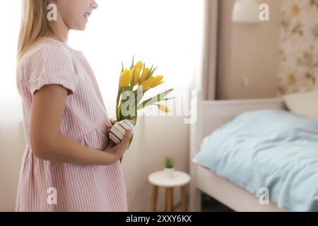 Osterkonzept. Kleines lächelndes Mädchen in weißen flauschigen Hasenohren und rosa Kleid mit gelben Tulpenblüten und bunten Eiern. Zimmereinrichtung. Stockfoto