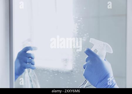 Verkürzte Ansicht der Haushälterin Hand in blauem Gummihandschuh, die chemische Reinigungsflüssigkeit in den Spiegel spritzt, während sie zu Hause das Badezimmer reinigt. Haushälterin Stockfoto