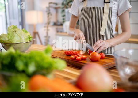 Ein zugeschnittenes Bild einer Frau in einer Schürze, die eine Tomate auf einem hölzernen Schneidebrett am Küchentisch schneidet und zu Hause kocht. Hausgemachtes Essen, gesundes Essen, Stockfoto