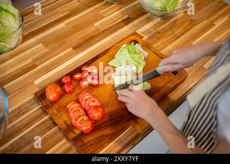 Ein Nahbild einer Frau, die Salat auf einem hölzernen Schneidebrett hackt, während sie Gemüse für ihren Salat am Küchentisch zubereitete, zuhause kocht. Stockfoto