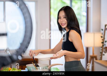 Eine attraktive asiatische Food Bloggerin kocht vor der Kamera und nimmt ein Video auf, wie sie in ihrer Küche gesundes Essen zubereitet. Stockfoto