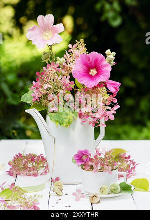 Blumenarrangement mit rosa, magentafarbenen Hollyhock- und lila Hortensie-Blüten in einer weißen Vintage Porzellan-Teekanne. (Alcea rosea) romantische Blumenstill Stockfoto