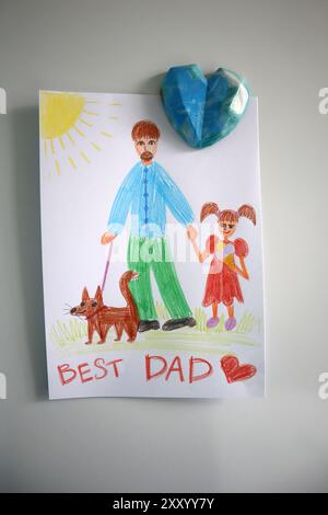 Fotopostkarte für den besten Vater, gezeichnet von einem Kind. Mit einem blauen Herzmagneten befestigt. Weißer Hintergrund. Zeichnen mit Farbstiften. Papa wird mit einem Mädchen und einem Hund dargestellt. Das Mädchen isst Eis. Stockfoto