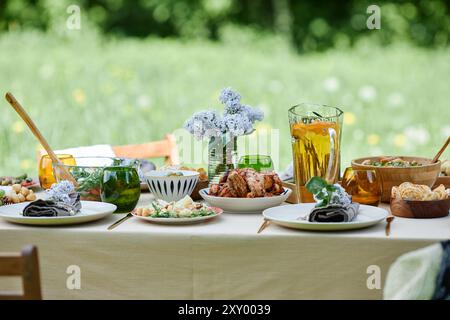 Esstisch im Freien mit verschiedenen Gerichten, frischen Blumen und erfrischenden Getränken. Schöner grüner Hintergrund schafft eine ruhige und einladende Atmosphäre Stockfoto