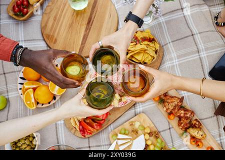 Gruppe von Freunden, die mit Getränken über dem Tisch mit verschiedenen Snacks und frischem Obst toasten, feiern im Freien an sonnigen Tagen und genießen gute Gesellschaft und köstliches Essen Stockfoto