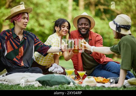 Gruppe von Freunden, die sich zum Picknick im Park versammeln, Gläser in temperamentvollem Toast hochheben, auf Picknickdecke sitzen, umgeben von der Natur, verschiedene Gruppen feiern gemeinsam Stockfoto