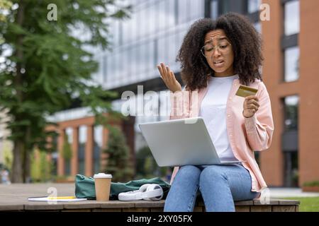 Verwirrte junge Frau mit Laptop und Kreditkarte im Freien. Frustration während der Online-Shopping-Transaktion auf der Campus-Bank zeigen. Bäume und modernes Gebäude im Hintergrund. Stockfoto