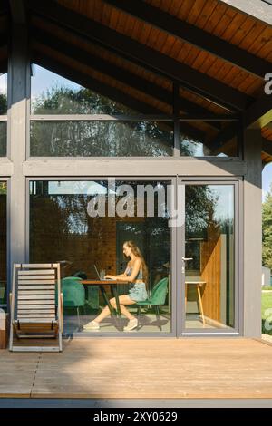 Frau arbeitet an einem Computer in einem modernen Landhaus mit einer großen Glasfassade an sonnigen Sommertagen Stockfoto