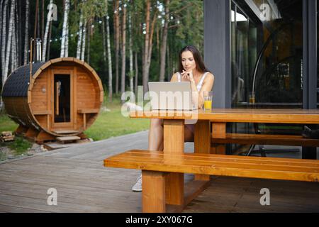 Entspannte Frau, die auf einem Laptop ein Video oder einen Film ansieht, während sie auf einer Holzbank auf der Terrasse eines Landhauses sitzt Stockfoto