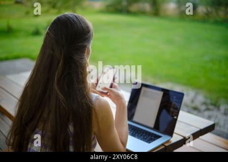 Eine Multitasking-Frau mit Laptop und Smartphone in ruhiger Gartenumgebung, während sie abends auf der Holzterrasse arbeitet Stockfoto