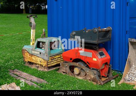 Ein paar unbenutzte Baumaschinen sitzen neben einem blauen Versandbehälter auf grünem Gras in einem Außenbereich. Stockfoto