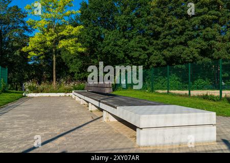 Eine elegante Betonbank befindet sich in einem friedlichen Park, im Sonnenlicht getaucht mit leuchtend grünen Bäumen im Hintergrund, die Besucher zum Entspannen und Genießen der Natur einladen. Stockfoto