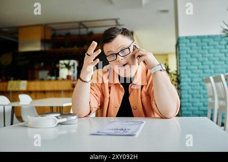 Ein Mann mit Down-Syndrom, mit Brille und Pfirsichjacke, sitzt an einem Kaffeetisch, spricht am Telefon und schaut sich die Speisekarte an. Stockfoto