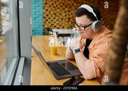 Ein Mann mit Down-Syndrom sitzt in einem Café, arbeitet an seinem Laptop, hat Kopfhörer an und einen Drink vor sich. Stockfoto