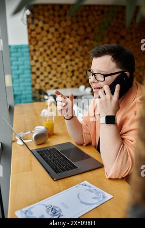 Ein Mann mit Down-Syndrom arbeitet ferngesteuert an seinem Laptop in einem Café und spricht mit seinem Telefon. Stockfoto