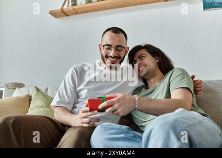 Zwei Männer teilen sich ein Lachen und ein Geschenk, während sie zusammen auf einer Couch sitzen. Stockfoto