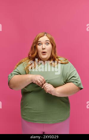 Eine Frau in grünem Hemd und rosa Hose starrt vor einem leuchtend rosa Hintergrund staunend. Stockfoto