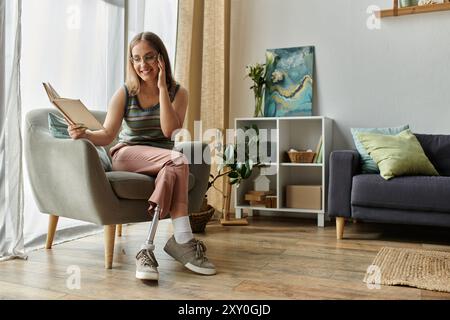 Eine junge Frau mit einer Beinprothese sitzt in einem bequemen Stuhl und liest in ihrem modernen Wohnzimmer ein Buch. Stockfoto