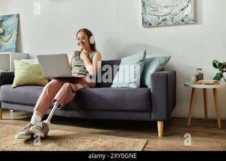 Eine junge Frau mit einer Beinprothese lächelt, während sie einen Laptop benutzt und Kopfhörer auf einer grauen Couch in ihrem modernen Wohnzimmer trägt. Stockfoto