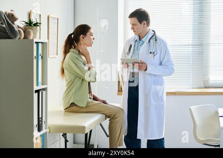 Arzt im Gespräch mit einer Patientin, die auf dem Untersuchungstisch in der Arztpraxis sitzt. Beide führten Gespräche, während sie Gesundheitsinformationen durchsahen Stockfoto